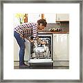 Woman Unloading Dish Washer In Kitchen. Framed Print