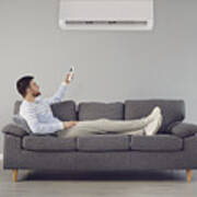 The Young Man Turns On The Air Conditioner Cools The Air While Sitting On The Sofa In The Room Art Print
