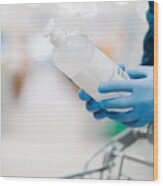 Unrecognizable Person In Medical Gloves Holds Bottle Of Sanitizer Gel, Poses In Shop, Leans At Shopping Cart, Buys Product For Coronavirus Protection, Blurred Background. Disinfection, Prevention Wood Print